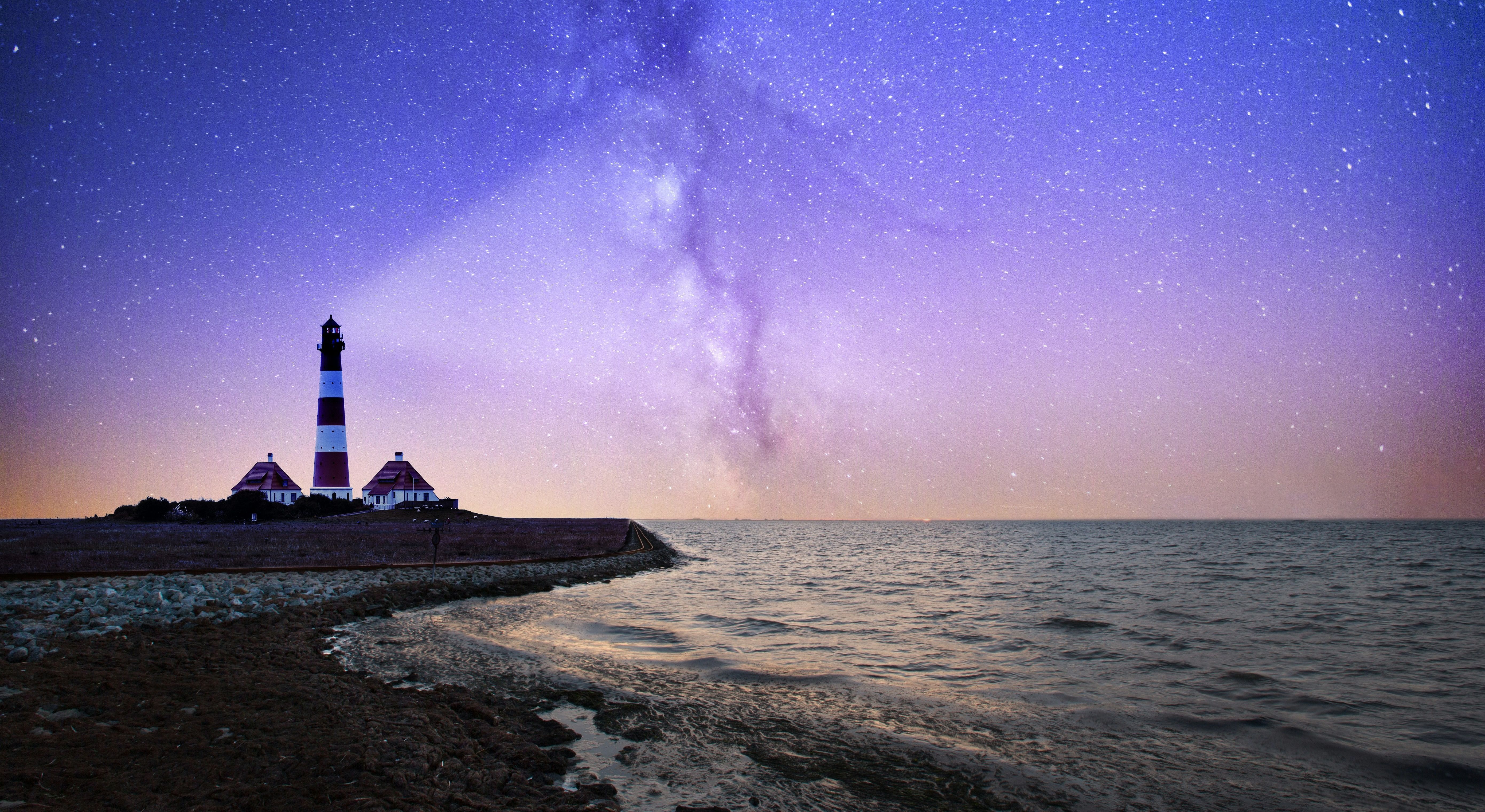 A red and white striped lighthouse shining out against the sea backdropped by a purple sky filled with nebula.