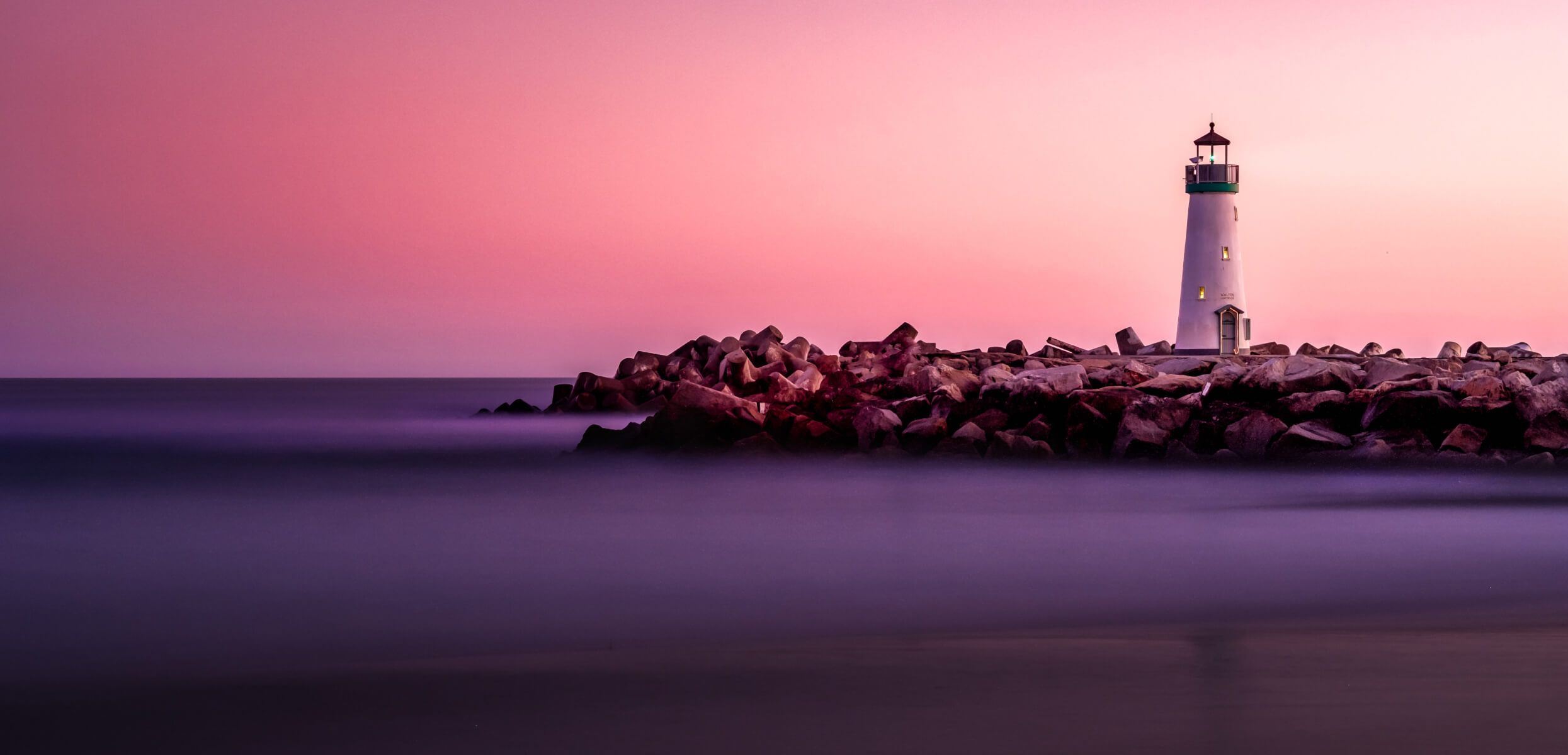 A lighthouse on a rocky shore, with purple water reflecting a rose sunset sky.